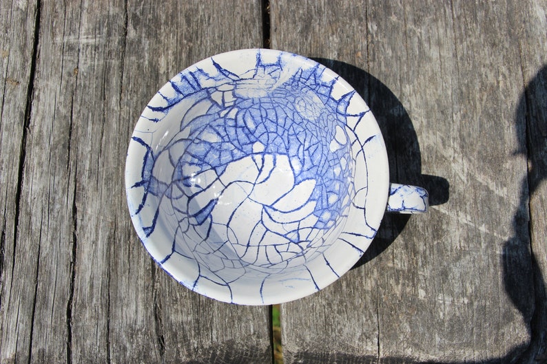 Puede incluir: Una taza de t&eacute; de cer&aacute;mica blanca con un esmalte craquelado azul. La taza tiene un asa y est&aacute; sentada sobre una superficie de madera.