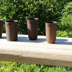 Könnte beinhalten: Fünf braune Keramikbecher mit roten Akzenten. Die Becher sind in einer Reihe auf einer Holzoberfläche angeordnet.
