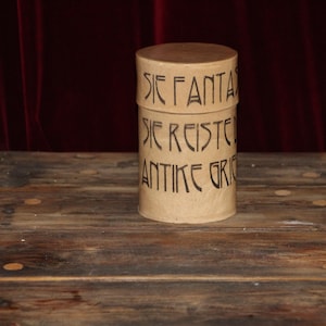 Puede incluir: Un recipiente cilíndrico de color beige con tapa, con texto negro: "SIE FANTA", "SIE REISTE" y "ANTIKER GRI". Se encuentra sobre una superficie de madera, con un fondo rojo oscuro.