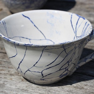 Puede incluir: Una taza de t&eacute; de cer&aacute;mica blanca con un esmalte craquelado azul. La taza tiene un asa y est&aacute; sentada sobre una superficie de madera.