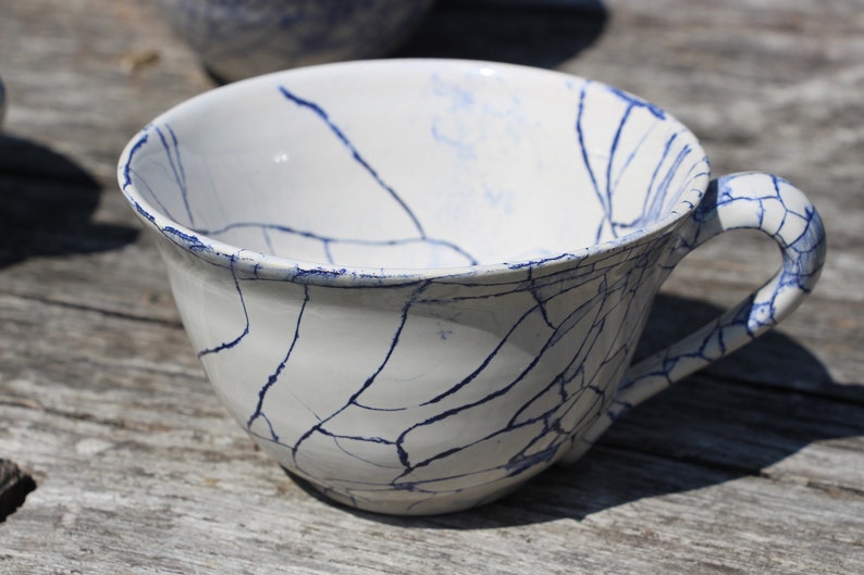 Puede incluir: Una taza de t&eacute; de cer&aacute;mica blanca con un esmalte craquelado azul. La taza tiene un asa y est&aacute; sentada sobre una superficie de madera.