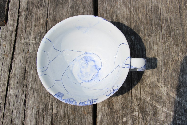 Puede incluir: Una taza de t&eacute; de cer&aacute;mica blanca con un dise&ntilde;o de remolino azul y blanco. La taza tiene un asa y est&aacute; sentada sobre una superficie de madera.