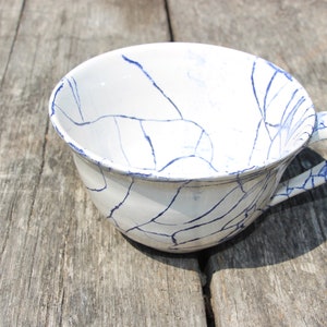 Puede incluir: Una taza de t&eacute; de cer&aacute;mica blanca con un dise&ntilde;o de craquelado azul. La taza tiene un asa y est&aacute; sentada sobre una superficie de madera.
