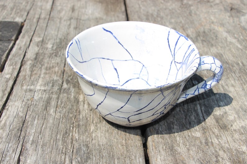 Puede incluir: Una taza de t&eacute; de cer&aacute;mica blanca con un dise&ntilde;o de craquelado azul. La taza tiene un asa y est&aacute; sentada sobre una superficie de madera.