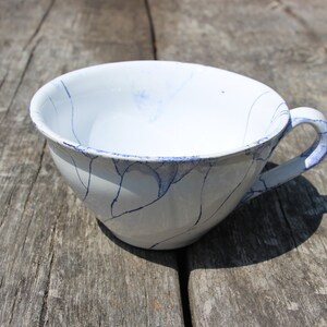 Puede incluir: Una taza de t&eacute; de cer&aacute;mica blanca con un dise&ntilde;o de craquelado azul y un asa. La taza est&aacute; sobre una superficie de madera.