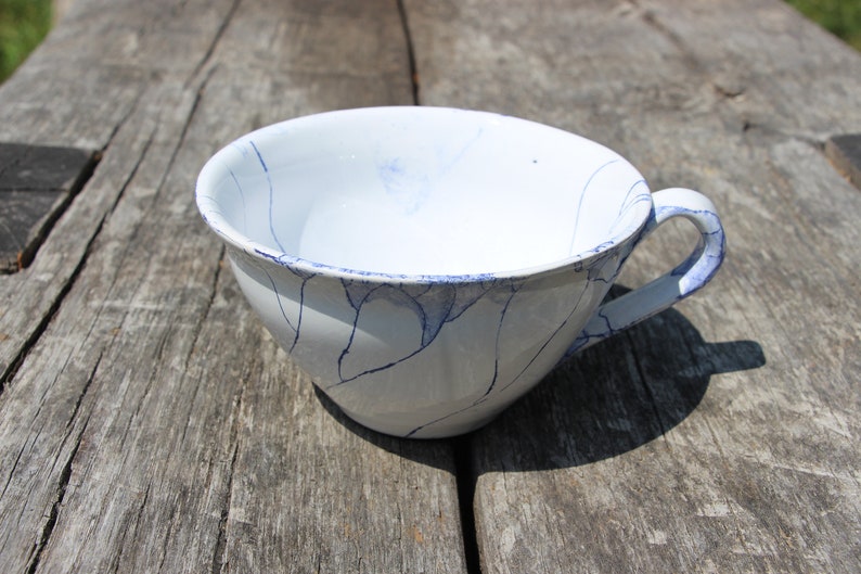 Puede incluir: Una taza de t&eacute; de cer&aacute;mica blanca con un dise&ntilde;o de craquelado azul y un asa. La taza est&aacute; sobre una superficie de madera.