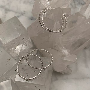 May include: Two pairs of silver hoop earrings. Each earring is made of small, evenly spaced beads. The earrings are displayed on a surface of clear, crystalline quartz.