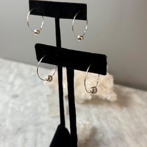 May include: Two pairs of silver hoop earrings displayed on a black stand. Each thin circular hoop features a single small, round, textured silver bead that slides along the wire. The metal has a bright, reflective finish throughout the design.