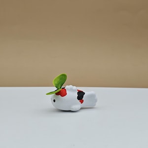 May include: A white ceramic figurine of a fish with red and black markings. The fish has a green leaf on its head.