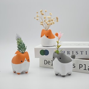 May include: Three ceramic cat-shaped planters in orange, gray, and white, each with a small opening for plants. The planters hold dried flowers and faux plants. The cat planters are on a white surface.