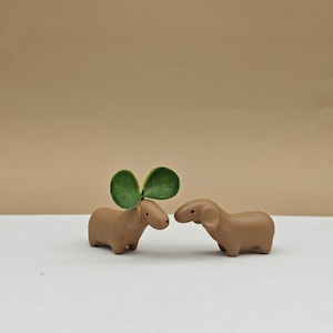 Puede incluir: Dos pequeñas figuritas de dachshund de cerámica marrón, una con una hoja verde en la cabeza, frente a frente sobre una superficie blanca.