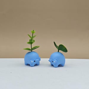 May include: Two small, light blue elephant-shaped planters with green plants sprouting from their heads. The planters have white dot eyes and small trunks. The background is a neutral tan color, and the planters are on a white surface.