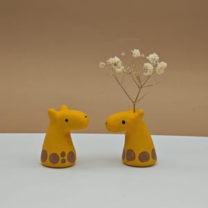 Puede incluir: Dos jarrones de cerámica amarillos en forma de jirafa con manchas marrones. Un jarrón tiene un ramo de flores blancas secas.
