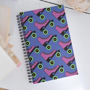 May include: A blue spiral notebook with a repeating pattern of pink roller skates with black wheels and green and black circles.