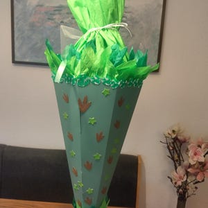 Op de afbeelding: Een kegelvormige schoolkegel in lichtgroen, versierd met bruine dinosaurusvoetafdrukken en kleine groene sterstickers. De bovenkant is gevuld met felgroen vloeipapier dat is samengebonden met een dun wit lint bij de opening.
