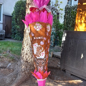 Könnte beinhalten: Eine große, kegelförmige Schultüte, verziert mit einem Leopardenmuster und dem Wort "NAME". Die Tüte ist braun mit rosa und orangefarbenem Seidenpapier oben und unten. Die Tüte ist mit rosa Blumen verziert.