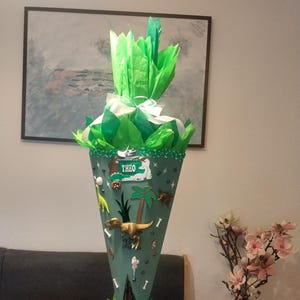 Op de afbeelding: Een kegelvormige schoolkegel in saliegroen, afgewerkt met felgroen vloeipapier en een wit lint. De kegel is versierd met dinosaurus-thema decoraties, waaronder een gouden T-Rex, palmbomen en een rechthoekig bordje met de naam THEO in het wit.