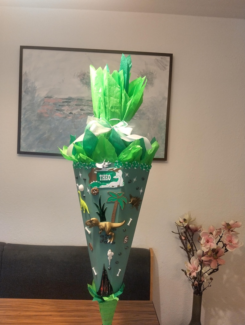 Op de afbeelding: Een kegelvormige schoolkegel in saliegroen, afgewerkt met felgroen vloeipapier en een wit lint. De kegel is versierd met dinosaurus-thema decoraties, waaronder een gouden T-Rex, palmbomen en een rechthoekig bordje met de naam THEO in het wit.
