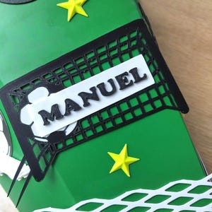 Peut inclure: Une bo&icirc;te cadeau verte d&eacute;cor&eacute;e sur le th&egrave;me du football. Elle pr&eacute;sente un filet de foot noir avec le nom "MANUEL" en lettres blanches, des &eacute;toiles jaunes et des d&eacute;coupes de ballons de foot. La bo&icirc;te est con&ccedil;ue pour un fan de foot.