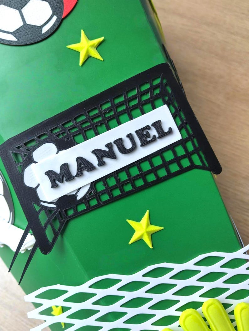 Peut inclure: Une bo&icirc;te cadeau verte d&eacute;cor&eacute;e sur le th&egrave;me du football. Elle pr&eacute;sente un filet de foot noir avec le nom "MANUEL" en lettres blanches, des &eacute;toiles jaunes et des d&eacute;coupes de ballons de foot. La bo&icirc;te est con&ccedil;ue pour un fan de foot.