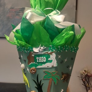 Op de afbeelding: Een kegelvormige schoolkegel versierd met dinosaurusfiguren, palmbomen en een naamplaatje met de tekst THEO. De bovenkant is gevuld met felgroen vloeipapier en gestrikt met een wit satijnen lint. De kegel bevat diverse kleine dinosaurus-versieringen.