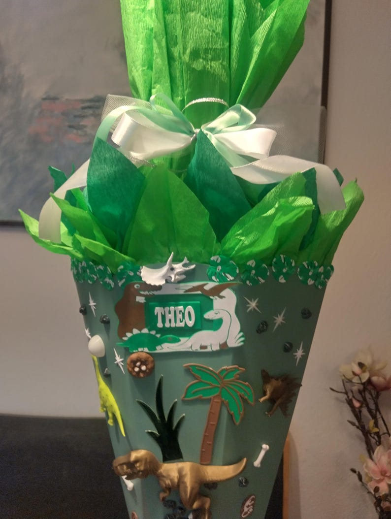 Op de afbeelding: Een kegelvormige schoolkegel versierd met dinosaurusfiguren, palmbomen en een naamplaatje met de tekst THEO. De bovenkant is gevuld met felgroen vloeipapier en gestrikt met een wit satijnen lint. De kegel bevat diverse kleine dinosaurus-versieringen.