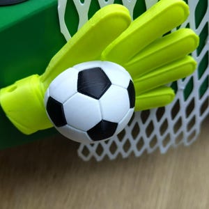 Peut inclure: Une petite cage de football avec un filet blanc, un gant de gardien de but vert fluo et un ballon de football noir et blanc. L'ensemble est pos&eacute; sur un fond vert, sugg&eacute;rant un th&egrave;me sportif.