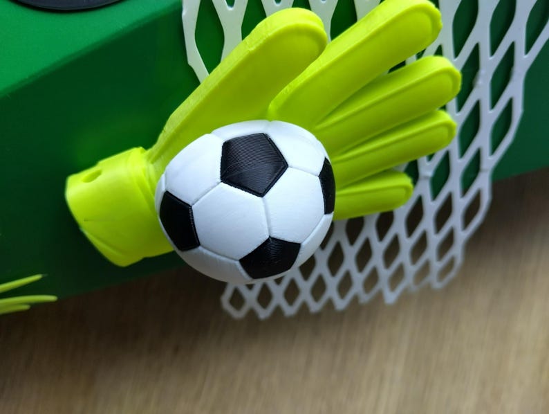 Peut inclure: Une petite cage de football avec un filet blanc, un gant de gardien de but vert fluo et un ballon de football noir et blanc. L'ensemble est pos&eacute; sur un fond vert, sugg&eacute;rant un th&egrave;me sportif.