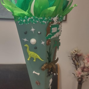 Op de afbeelding: Een saliegroene kegelvormige geschenkzak, afgewerkt met felgroen vloeipapier en witte linten. De kegel is versierd met diverse dinosaurusfiguren, witte botvormen, steraccenten en een rechthoekig bordje met de naam THEO in het wit.