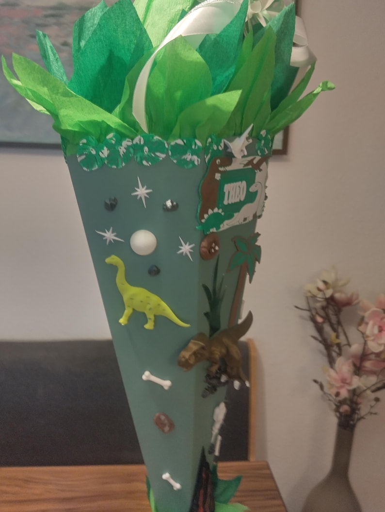 Op de afbeelding: Een saliegroene kegelvormige geschenkzak, afgewerkt met felgroen vloeipapier en witte linten. De kegel is versierd met diverse dinosaurusfiguren, witte botvormen, steraccenten en een rechthoekig bordje met de naam THEO in het wit.