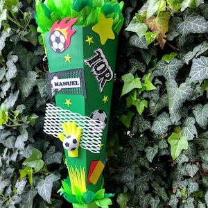 Peut inclure: Un cornet scolaire vert en forme de c&ocirc;ne, d&eacute;cor&eacute; sur le th&egrave;me du football. Il pr&eacute;sente un ballon de football, un gant de gardien et le nom "MANUEL". Le sac est orn&eacute; d'&eacute;toiles, de flammes et du mot "TOP". Le sac est rempli de papier de soie vert et jaune.