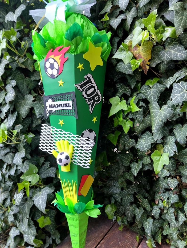 Peut inclure: Un cornet scolaire vert en forme de c&ocirc;ne, d&eacute;cor&eacute; sur le th&egrave;me du football. Il pr&eacute;sente un ballon de football, un gant de gardien et le nom "MANUEL". Le sac est orn&eacute; d'&eacute;toiles, de flammes et du mot "TOP". Le sac est rempli de papier de soie vert et jaune.