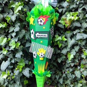 Peut inclure: Un cornet cadeau vert en forme de c&ocirc;ne, d&eacute;cor&eacute; sur le th&egrave;me du football. Il pr&eacute;sente des ballons de football, des &eacute;toiles et le nom "MANUEL". Le haut est rempli de papier de soie vert et nou&eacute; avec un ruban blanc. Le tout est pos&eacute; sur un fond de feuillage vert.