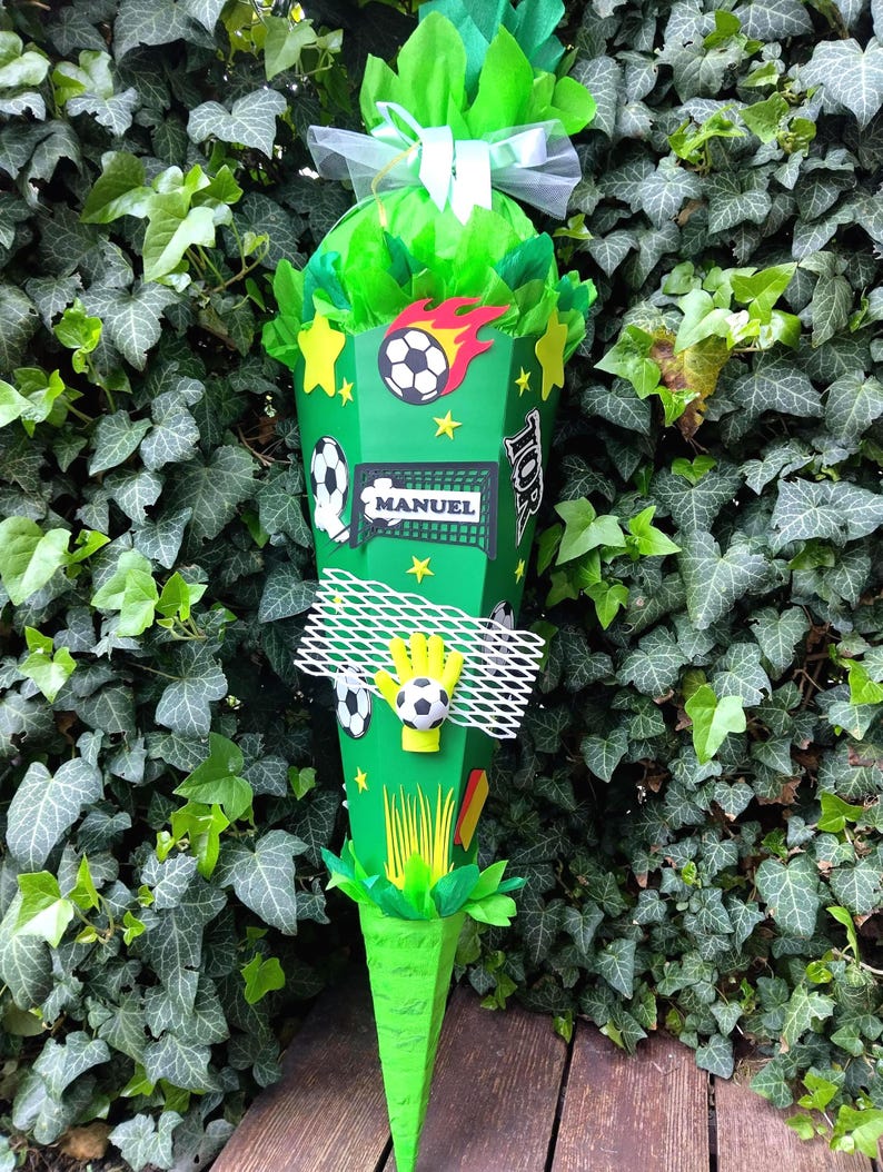 Peut inclure: Un cornet cadeau vert en forme de c&ocirc;ne, d&eacute;cor&eacute; sur le th&egrave;me du football. Il pr&eacute;sente des ballons de football, des &eacute;toiles et le nom "MANUEL". Le haut est rempli de papier de soie vert et nou&eacute; avec un ruban blanc. Le tout est pos&eacute; sur un fond de feuillage vert.