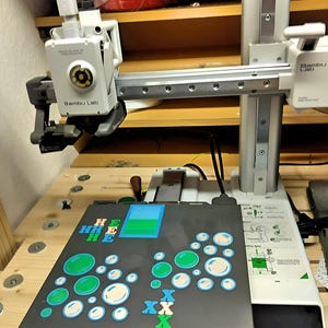Peut inclure: Une imprimante 3D avec un cadre gris et blanc, portant le logo "Bambu Lab". L'imprimante est install&eacute;e sur une surface en bois, avec un plateau d'impression noir et gris affichant des formes color&eacute;es et le texte "HEE".
