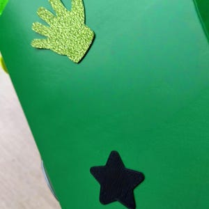 Peut inclure: Une surface vert vif avec une d&eacute;coupe en forme de main verte scintillante et une d&eacute;coupe en forme d'&eacute;toile noire. La d&eacute;coupe de la main a un aspect textur&eacute; et scintillant. L'&eacute;toile est noire unie.