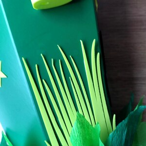 Peut inclure: Gros plan sur une bo&icirc;te verte d&eacute;cor&eacute;e d'herbe et de feuilles jaune-vert d&eacute;coup&eacute;es. Un objet cylindrique vert clair est visible en haut. La bo&icirc;te a une d&eacute;coupe en forme d'&eacute;toile sur le c&ocirc;t&eacute;.