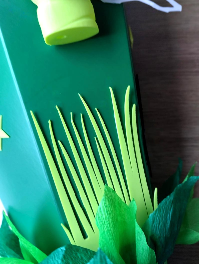 Peut inclure: Gros plan sur une bo&icirc;te verte d&eacute;cor&eacute;e d'herbe et de feuilles jaune-vert d&eacute;coup&eacute;es. Un objet cylindrique vert clair est visible en haut. La bo&icirc;te a une d&eacute;coupe en forme d'&eacute;toile sur le c&ocirc;t&eacute;.