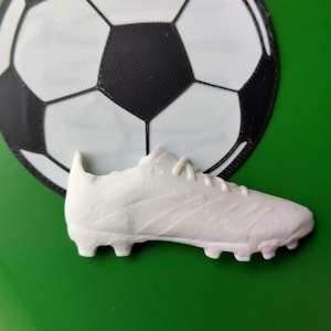 Peut inclure: Un crampon de football blanc est plac&eacute; devant un graphisme de ballon de football noir et blanc sur fond vert. Le crampon a une surface textur&eacute;e et des crampons sur la semelle.