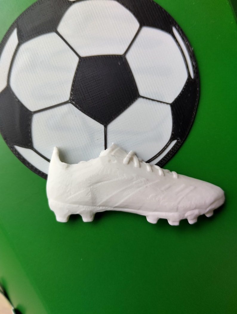 Peut inclure: Un crampon de football blanc est plac&eacute; devant un graphisme de ballon de football noir et blanc sur fond vert. Le crampon a une surface textur&eacute;e et des crampons sur la semelle.