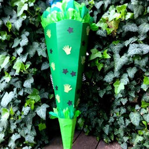 Peut inclure: Un cornet de bonbons vert en forme de c&ocirc;ne, d&eacute;cor&eacute; d'empreintes de mains jaunes et d'&eacute;toiles noires. Le sac est rempli de papier de soie vert et bleu, devant un fond de lierre.