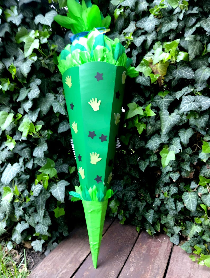 Peut inclure: Un cornet de bonbons vert en forme de c&ocirc;ne, d&eacute;cor&eacute; d'empreintes de mains jaunes et d'&eacute;toiles noires. Le sac est rempli de papier de soie vert et bleu, devant un fond de lierre.