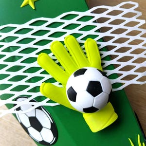 Peut inclure: Une bo&icirc;te d&eacute;corative verte sur le th&egrave;me du football. Elle pr&eacute;sente un gant de gardien de but jaune tenant un ballon de football noir et blanc, un filet blanc et une &eacute;toile jaune. Un graphisme de ballon de football est &eacute;galement visible.
