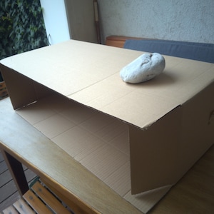 Op de afbeelding: Een rechthoekige bruine kartonnen structuur met een open voorzijde en een glad bovenoppervlak. Een enkele, onregelmatige, lichtgrijze steen met een getextureerd oppervlak is geplaatst op de rechterbovenhoek van de kartonnen doos.