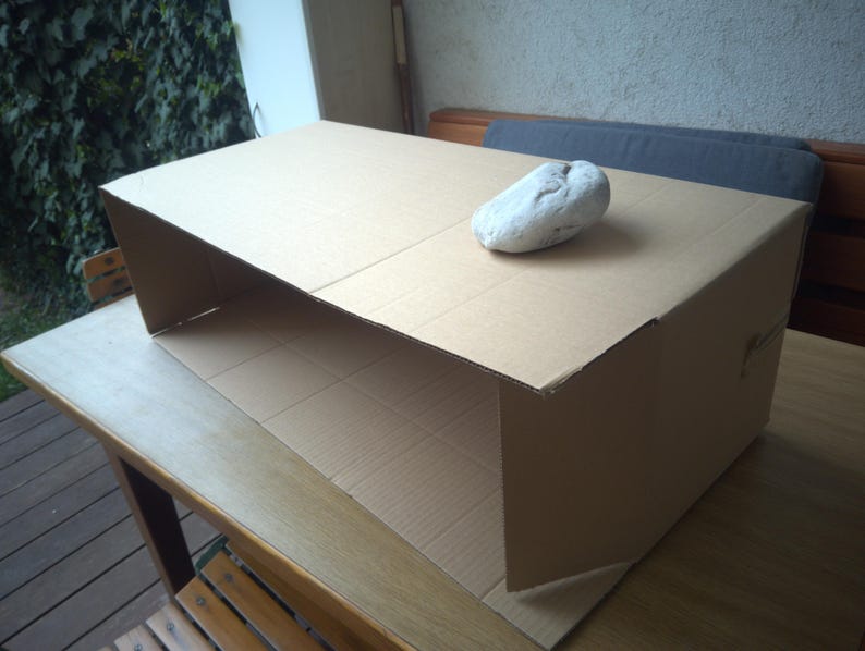 Op de afbeelding: Een rechthoekige bruine kartonnen structuur met een open voorzijde en een glad bovenoppervlak. Een enkele, onregelmatige, lichtgrijze steen met een getextureerd oppervlak is geplaatst op de rechterbovenhoek van de kartonnen doos.