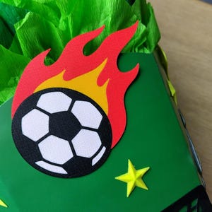 Peut inclure: Un coffret cadeau vert d&eacute;cor&eacute; d'un ballon de football en flammes et d'&eacute;toiles jaunes. Le ballon de football est noir et blanc. Les flammes sont rouges et jaunes. Le fond est en papier de soie vert.