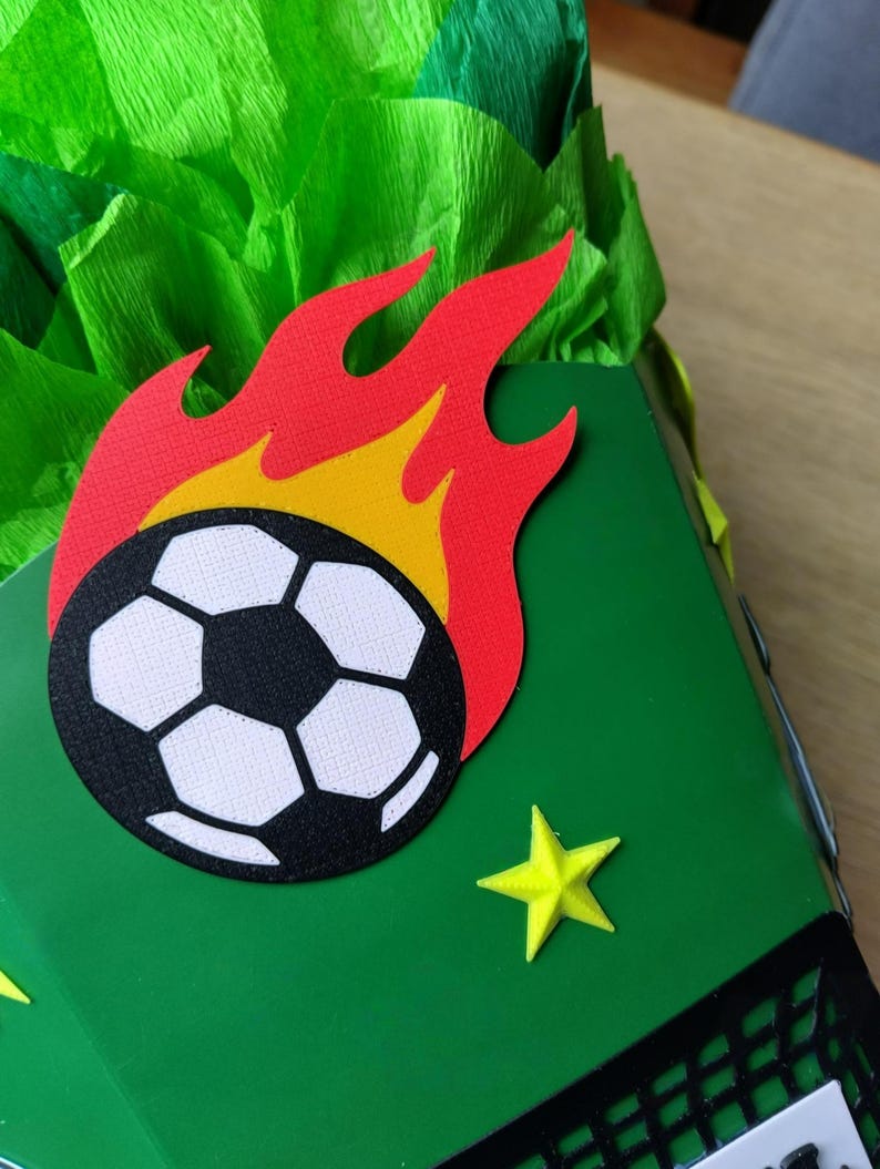 Peut inclure: Un coffret cadeau vert d&eacute;cor&eacute; d'un ballon de football en flammes et d'&eacute;toiles jaunes. Le ballon de football est noir et blanc. Les flammes sont rouges et jaunes. Le fond est en papier de soie vert.