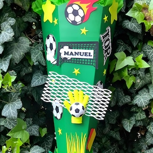 Peut inclure: Une bo&icirc;te cadeau verte en forme de c&ocirc;ne d&eacute;cor&eacute;e de d&eacute;coupes sur le th&egrave;me du football. La bo&icirc;te pr&eacute;sente des ballons de football, des &eacute;toiles, un but de football avec le nom "MANUEL" et un gant de gardien tenant un ballon. Le dessus est rempli de papier de soie vert.