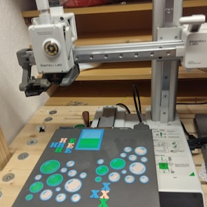 Op de afbeelding: Een witte Bambu Lab 3D-printer met een printkop boven een zwarte bouwplaat. De plaat bevat geprinte ontwerpen, waaronder groene en witte cirkelvormige bubbelpatronen, blauwe en oranje letter H- en X-vormen, en een blauw en groen rechthoek.