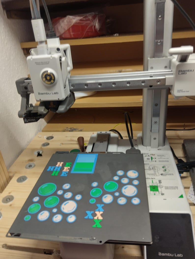 Op de afbeelding: Een witte Bambu Lab 3D-printer met een printkop boven een zwarte bouwplaat. De plaat bevat geprinte ontwerpen, waaronder groene en witte cirkelvormige bubbelpatronen, blauwe en oranje letter H- en X-vormen, en een blauw en groen rechthoek.